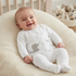 A smiling baby wearing a white long-sleeve footed sleeper with a gray elephant graphic, lying on a cream-colored textured baby lounger in a nursery setting.