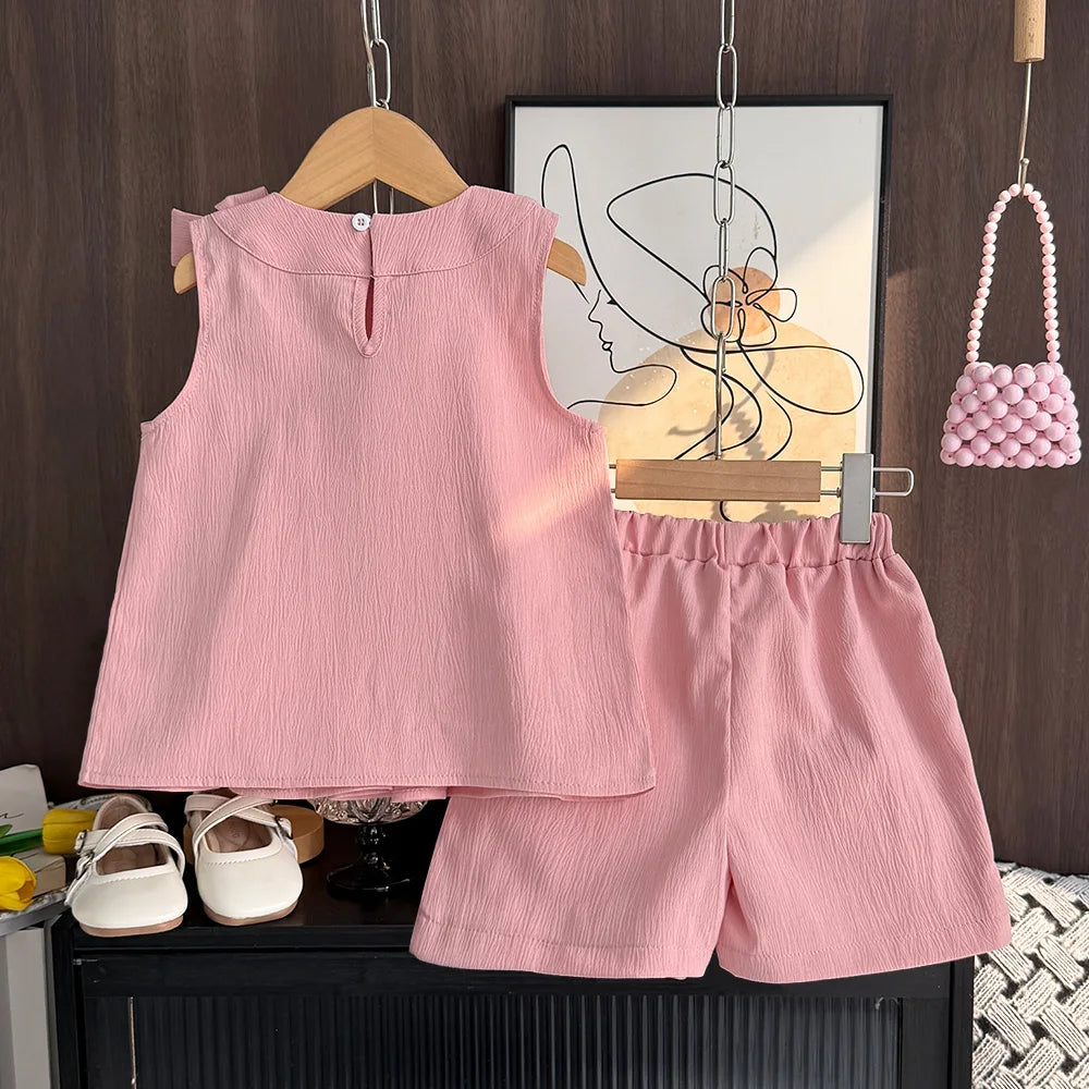 Pink children's outfit with shorts and a top on a hanger, with a small pink handbag and white shoes in the background.