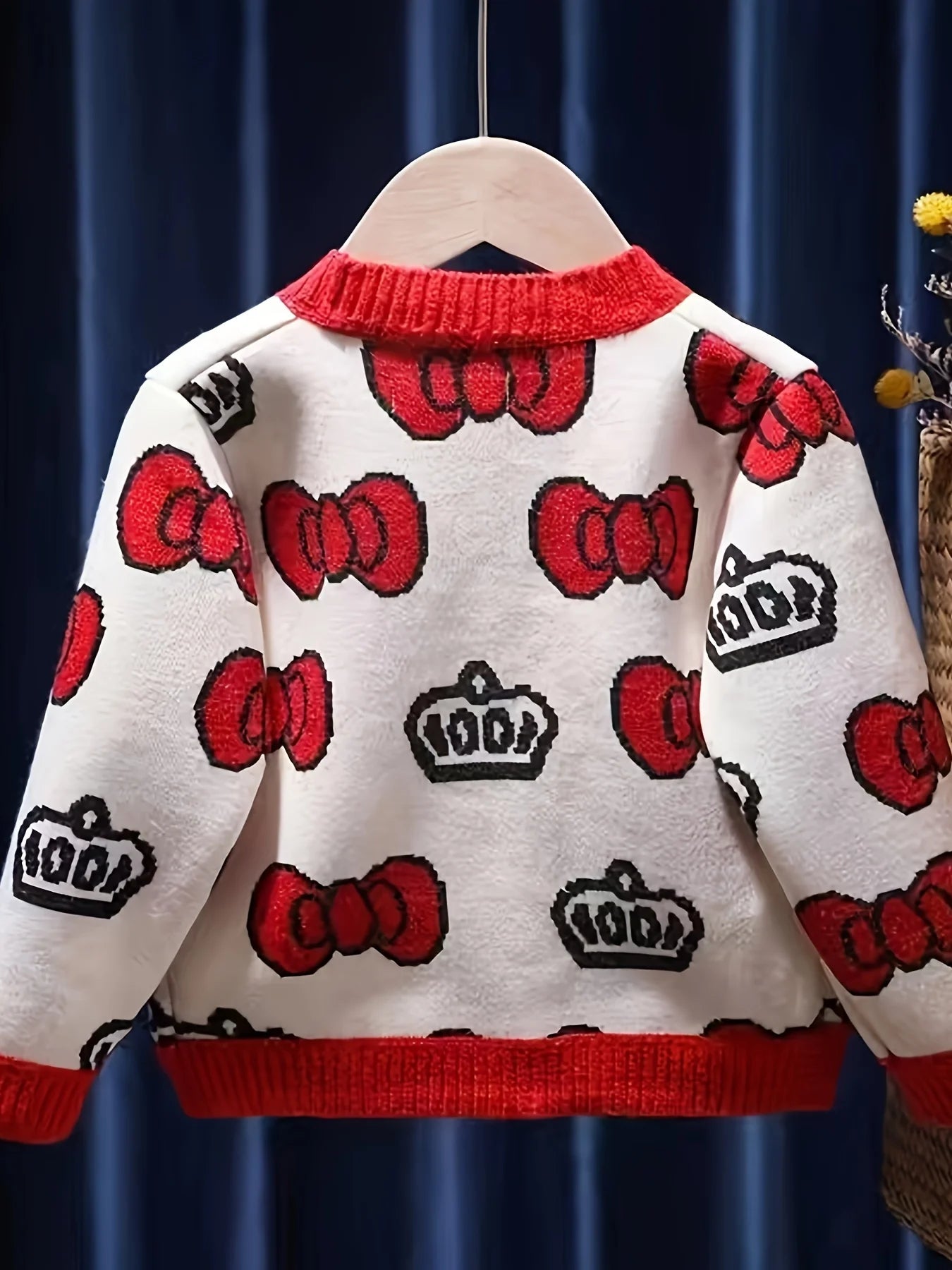 Children's sweater with red bows and crowns on a hanger against a dark curtain background