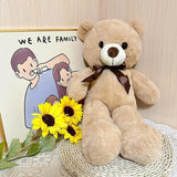 Large teddy bear with a bow tie, sunflowers, and a small sign reading 'We Are Family' on a light wooden background.