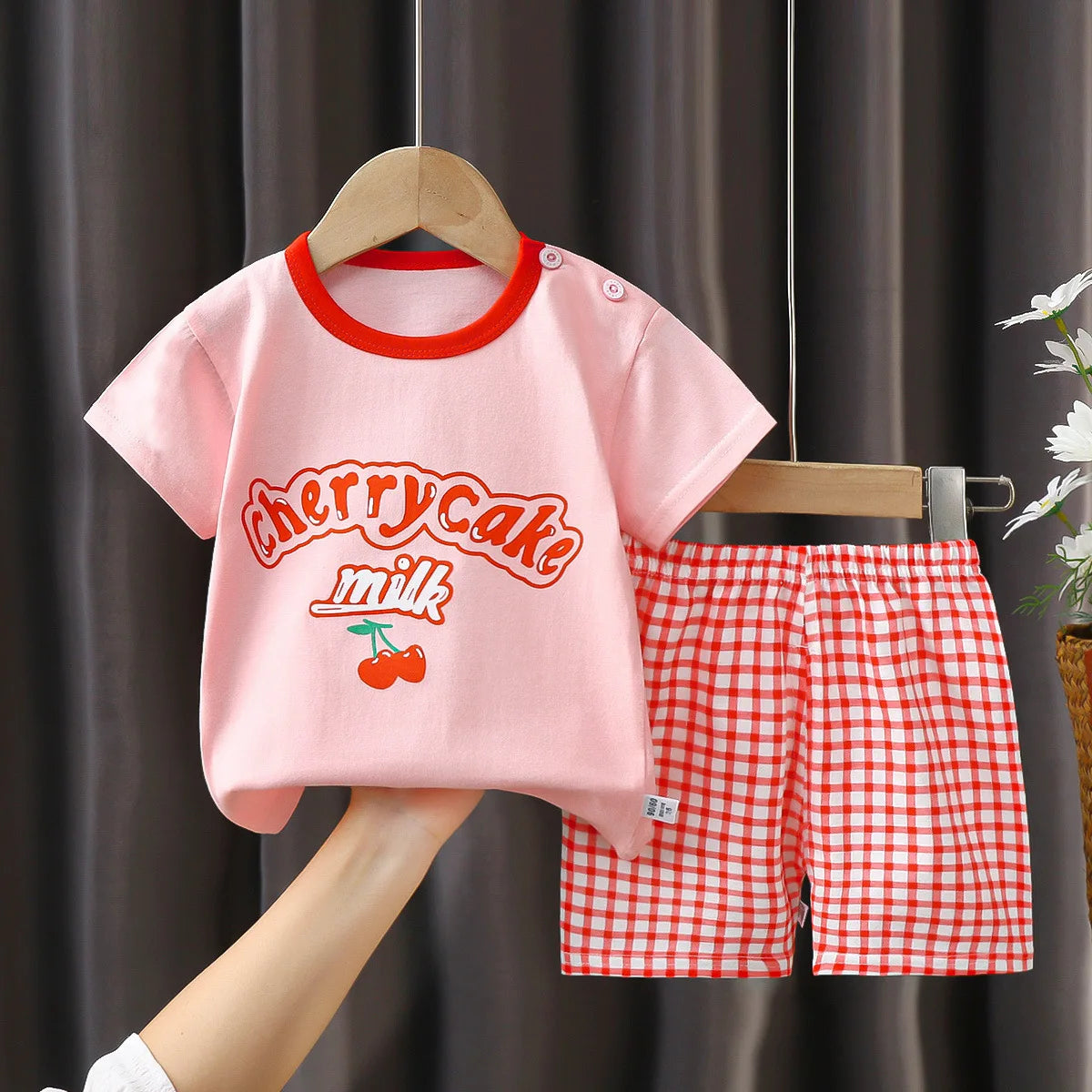 Children's pink t-shirt with red trim and checkered shorts on a hanger against a dark curtain background.