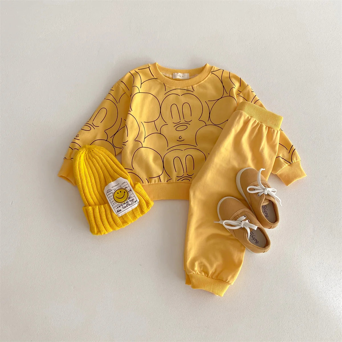 Yellow children's outfit with Mickey Mouse design, hat, and shoes on a light gray background