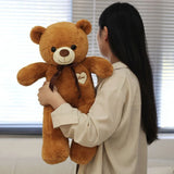 Person holding a large brown teddy bear with a heart ribbon in front of a window.