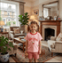 Young girl in a living room wearing a pink 'cherry cake' shirt and red checkered shorts, holding a teddy bear.