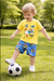 A young child in a yellow cartoon T-shirt and blue shorts kicks a black-and-white soccer ball while playing on green grass in a sunny park with trees in the background