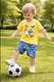 A young child in a yellow cartoon T-shirt and blue shorts kicks a black-and-white soccer ball while playing on green grass in a sunny park with trees in the background