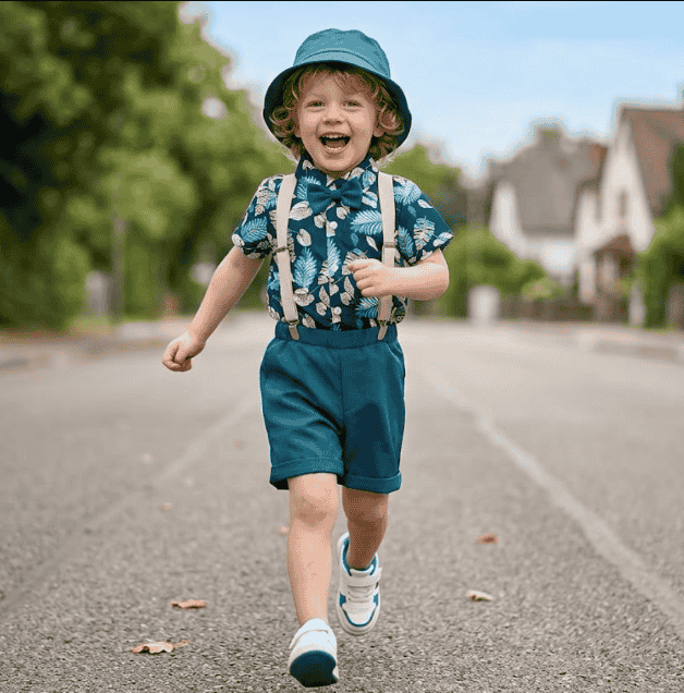 Child running on a street wearing a blue hat, floral shirt, suspenders, and shorts.