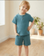 Child wearing a teal outfit standing in a room with wooden floor and plants.