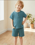 Child wearing a teal outfit standing in a room with wooden floor and plants.