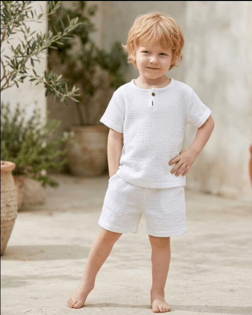 Young child in a white outfit standing outdoors with plants in the background