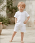 Young child in a white outfit standing outdoors with plants in the background