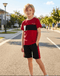 Young boy in a red and black sports outfit standing on a street.