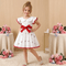 Young girl in a white dress with red accents standing in front of a beige wall with floral decorations.