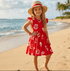 Young girl in a red dress with white floral patterns and a straw hat standing on a beach.