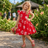 Young girl in a red dress with white floral patterns standing outdoors among plants.