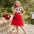 Young girl in a red and white dress standing outdoors with flowers and a bench in the background.