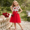 Young girl in a red and white dress standing outdoors with flowers and a bench in the background.