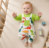 Baby wearing a green shirt and dinosaur-patterned overalls lying on a beige surface with toys around.
