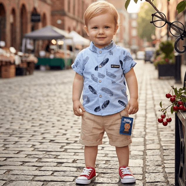 Child wearing a blue shirt with feather pattern and beige shorts on a cobblestone street.