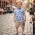 Child wearing a blue shirt with feather pattern and beige shorts on a cobblestone street.