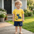 Child wearing a yellow shirt with a dinosaur design and blue shorts standing on a wooden deck.