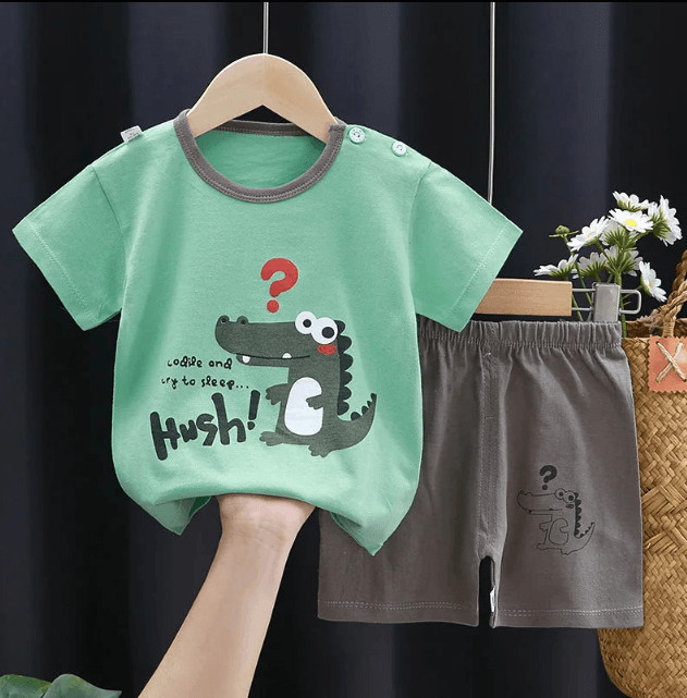 Children's green t-shirt with a crocodile graphic and gray shorts on a hanger against a dark background.