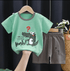Children's green t-shirt with a crocodile graphic and gray shorts on a hanger against a dark background.