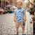 Child wearing a blue shirt with feather pattern and beige shorts on a cobblestone street.