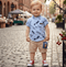 Child wearing a blue shirt with feather pattern and beige shorts on a cobblestone street.
