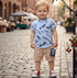 Child wearing a blue shirt with feather pattern and beige shorts on a cobblestone street.