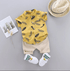 Children's outfit with yellow shirt, beige shorts, white shoes, and straw hat on a gray background.