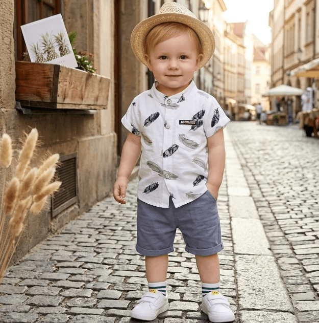 Child wearing a white shirt with feather pattern and blue shorts on a cobblestone street.