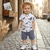 Child wearing a white shirt with feather pattern and blue shorts on a cobblestone street.
