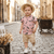 Child wearing a pink shirt with black patterns and beige shorts standing on a cobblestone street.