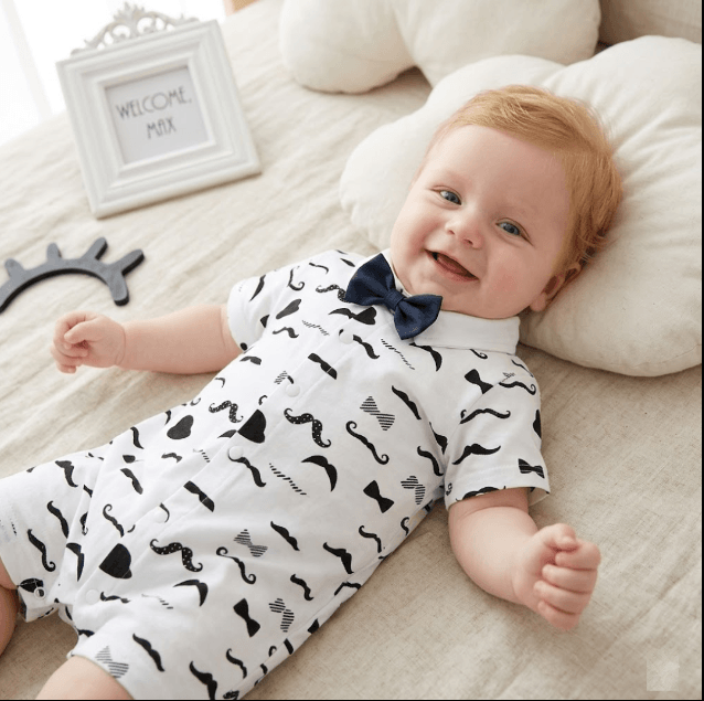 Baby wearing a white outfit with black patterns and a bow tie, lying on a bed.