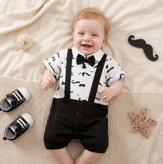 Baby wearing a formal outfit with suspenders and a bow tie, surrounded by decorative items on a beige blanket.