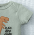 Light green baby onesie with brown dinosaur and black text 'little dino dude' on a textured white background