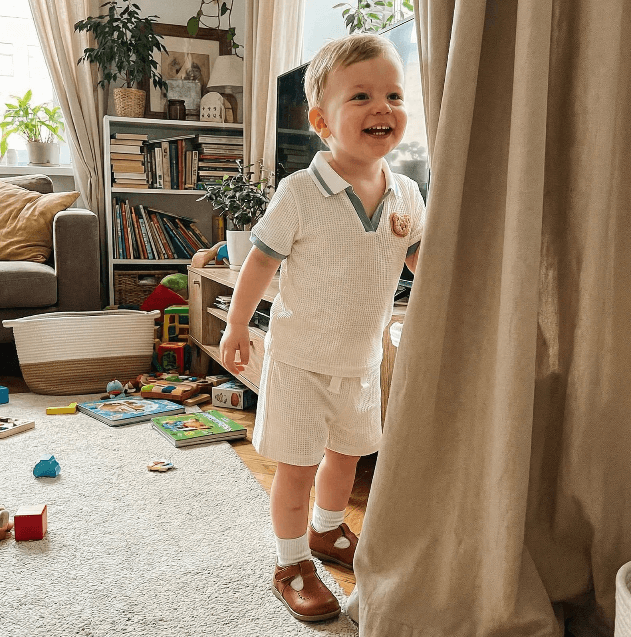 Child in a white outfit standing in a living room with toys on the floor.