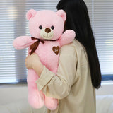 Person holding a large pink teddy bear with a heart on a blurred indoor background