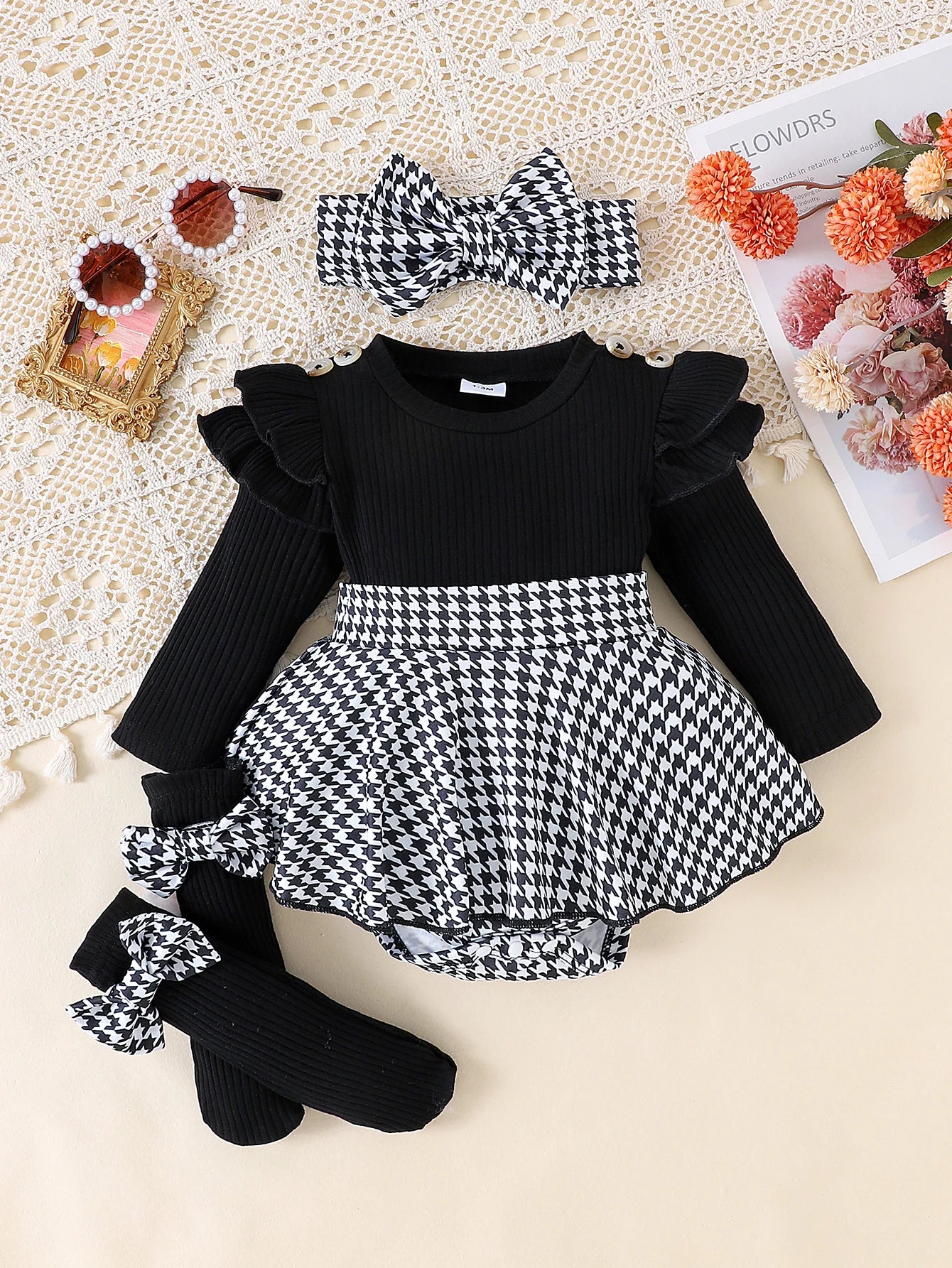 Black and white baby outfit with ruffled long-sleeve top, houndstooth skirt, socks, and headband on a textured surface.