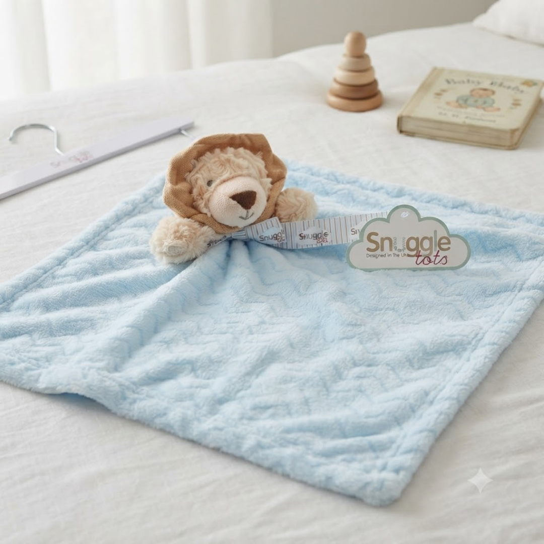 A light blue textured baby lovey blanket with a plush lion head attached, styled on a white bed with a wooden hanger, a wooden stacking toy, and a book in the background. A 'Snuggle tots' brand tag is visible.