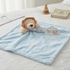 A light blue textured baby lovey blanket with a plush lion head attached, styled on a white bed with a wooden hanger, a wooden stacking toy, and a book in the background. A 'Snuggle tots' brand tag is visible.