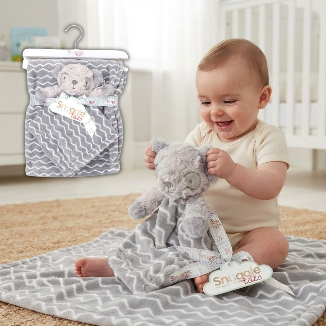 A baby sitting on a blanket in a nursery holds a plush gray teddy bear wrapped in a gray and white chevron Snuggle Baby blanket. An inset in the upper left shows the packaged product on a hanger.
