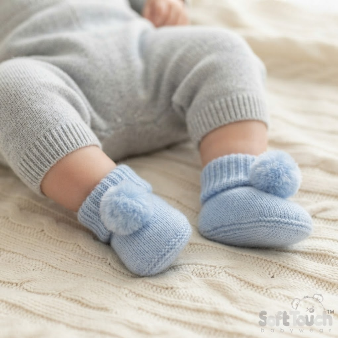 A close-up of a baby's legs wearing light blue knit booties with fluffy pom-poms, resting on a cream-colored cable-knit blanket. The baby is also wearing a light gray knit outfit.