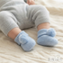 A close-up of a baby's legs wearing light blue knit booties with fluffy pom-poms, resting on a cream-colored cable-knit blanket. The baby is also wearing a light gray knit outfit.
