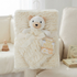 A cream-colored textured blanket with a plush teddy bear attached at the top, tied with a matching ribbon, and still on its retail tag. The blanket is displayed on a hanger in a soft, neutral nursery setting with a sofa and crib in the background.
