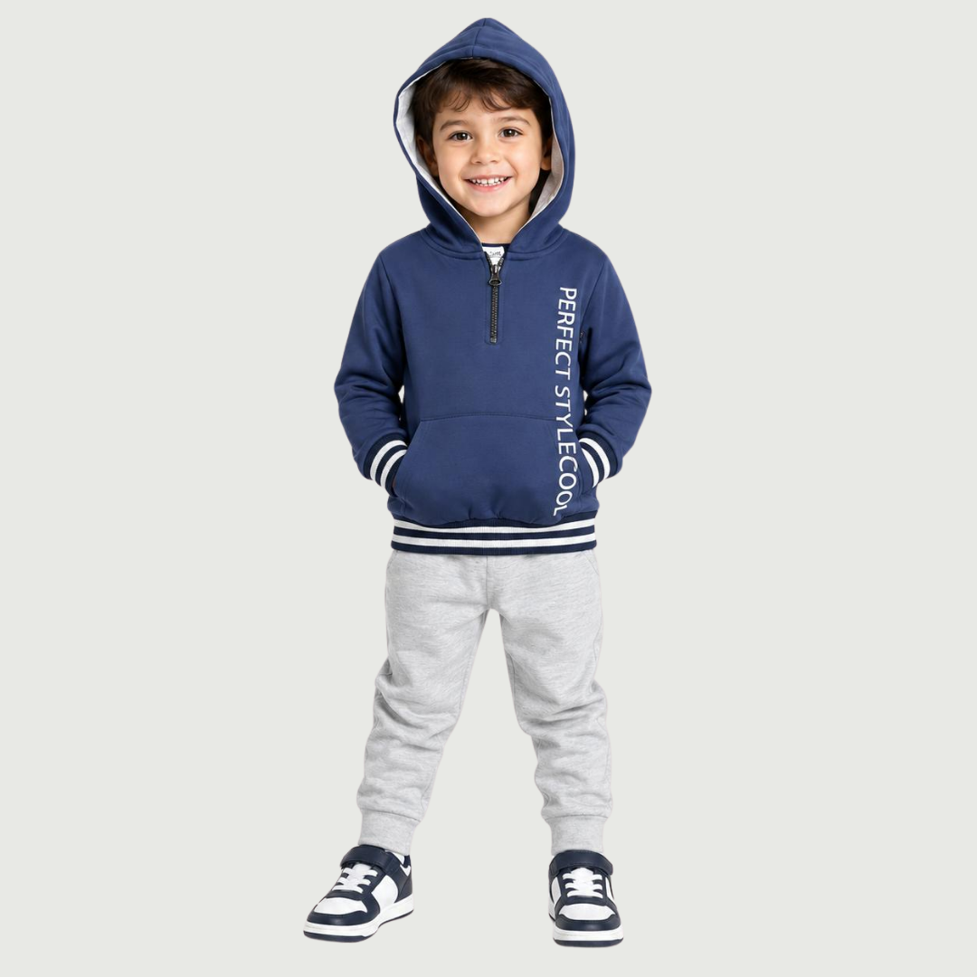 A smiling toddler standing against a plain white background, wearing a cream hooded zip jacket with a blue and beige animal print, a white graphic T-shirt, light blue pants, and white sneakers.