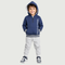 A smiling toddler standing against a plain white background, wearing a cream hooded zip jacket with a blue and beige animal print, a white graphic T-shirt, light blue pants, and white sneakers.