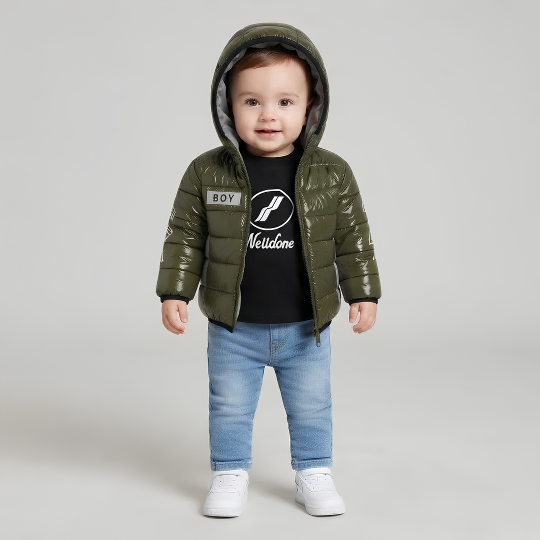 A toddler standing against a light gray studio background wearing a shiny olive green hooded puffer jacket over a black graphic T-shirt, blue jeans, and white sneakers. The jacket has a visible 'BOY Meuloone' patch on the chest.