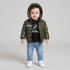 A toddler standing against a light gray studio background wearing a shiny olive green hooded puffer jacket over a black graphic T-shirt, blue jeans, and white sneakers. The jacket has a visible 'BOY Meuloone' patch on the chest.